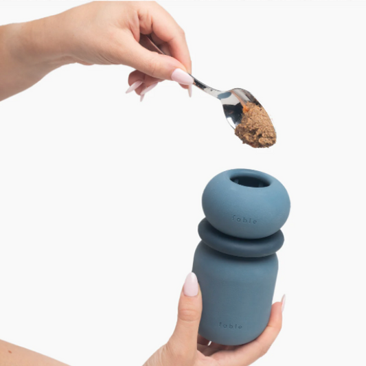 Blue silicone container with a spoonful of brown powder, held by a hand against a white background.